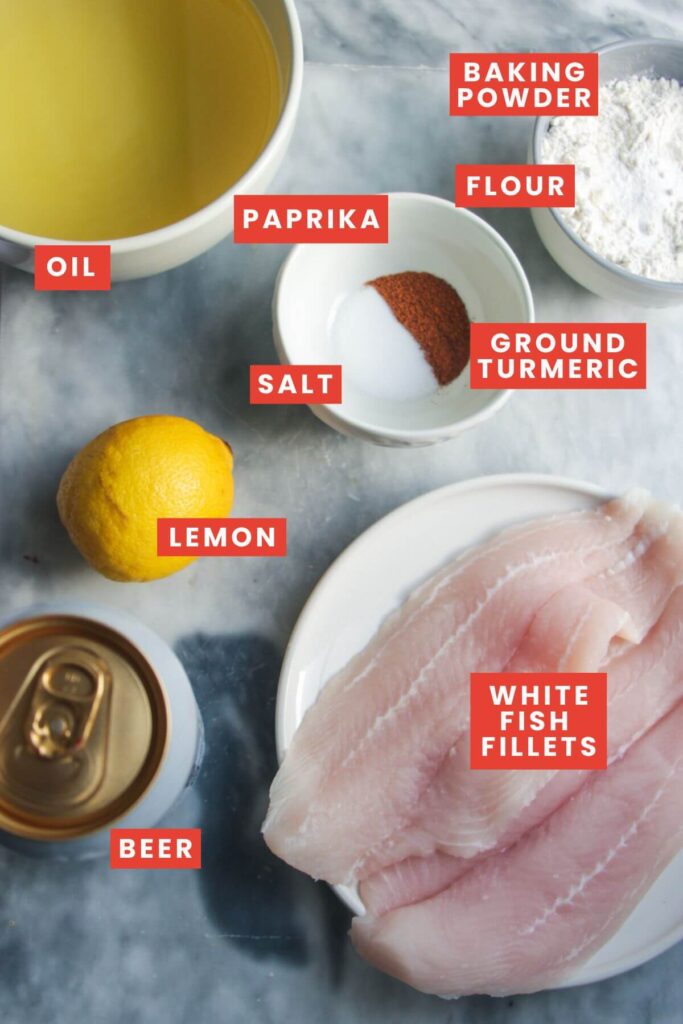 Ingredients for beer battered fish laid out on a grey marble background and labelled.