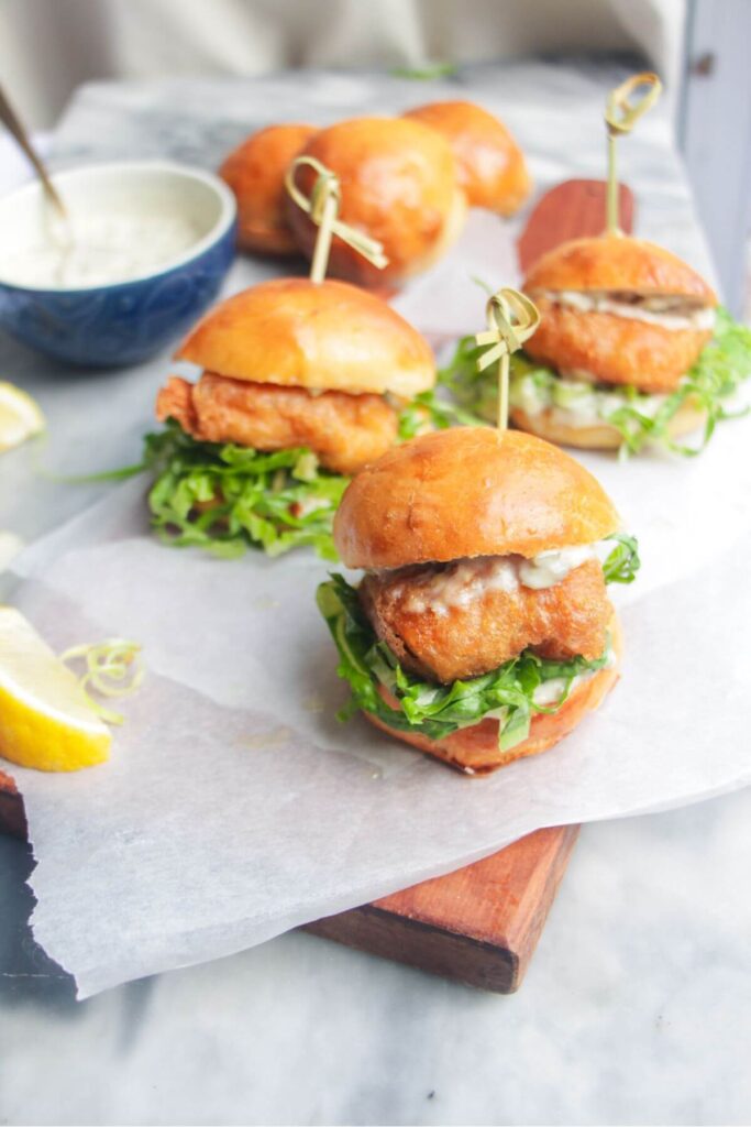 Three beer battered fish sliders on a baking paper lined wooden board with tartare sauce on the side.