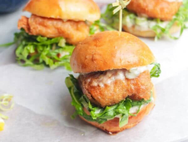 Three beer battered fish sliders on a baking paper lined wooden board with tartare sauce on the side.