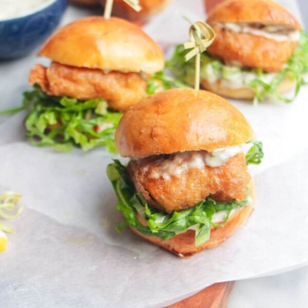 Three beer battered fish sliders on a baking paper lined wooden board with tartare sauce on the side.