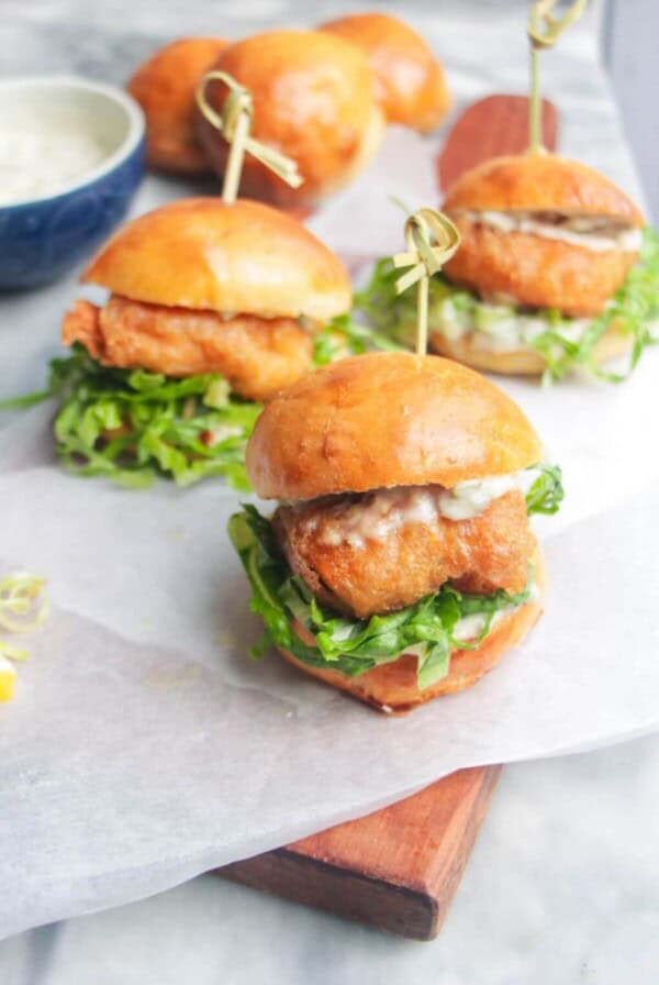 Three beer battered fish sliders on a baking paper lined wooden board with tartare sauce on the side.
