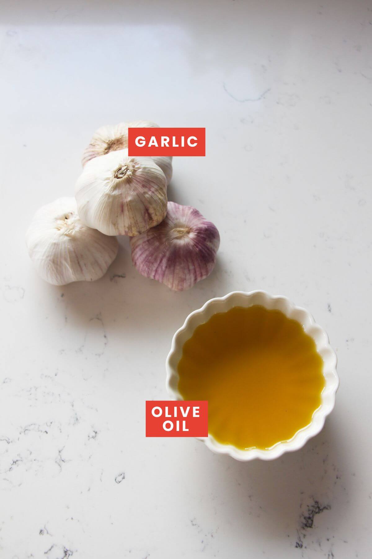 Ingredients for confit garlic laid out on a white marble background and labelled.