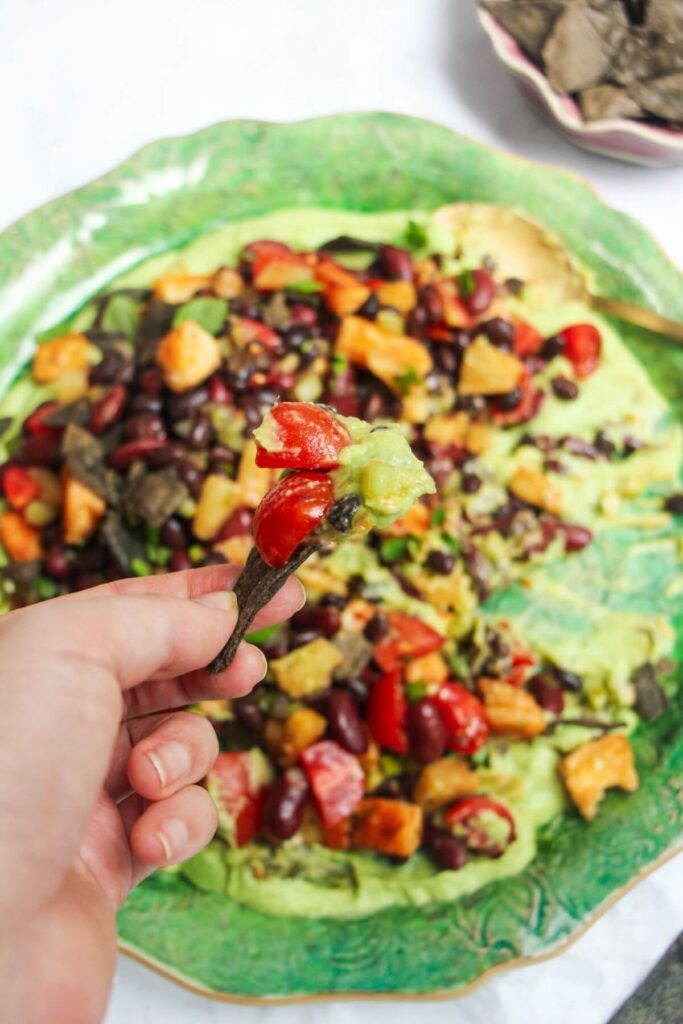 Hand holding up a blue corn tortilla chip with whipped avocado and cherry tomatoes on it.
