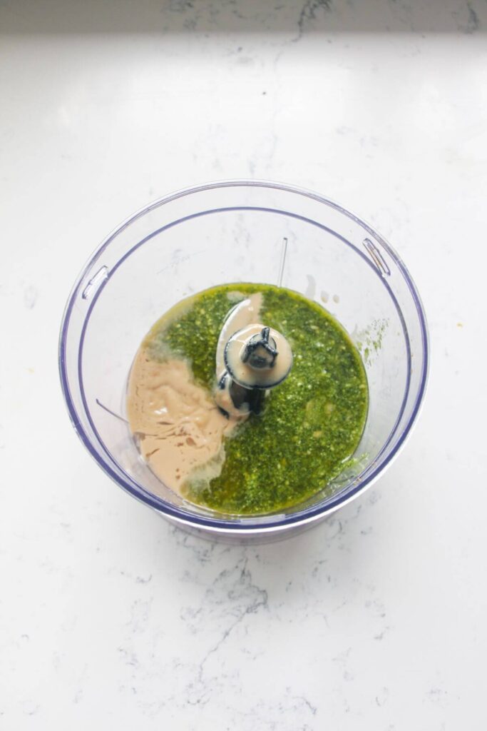 Tahini and pesto in a small food chopper on a white marble background.