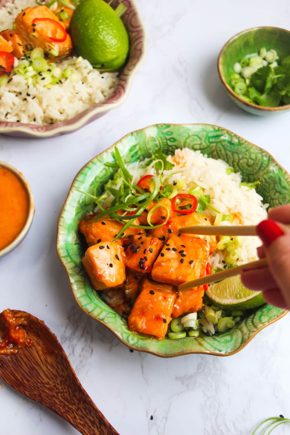 Hand using chopsticks about to pick up a piece of bang bang salmon in a bowl.