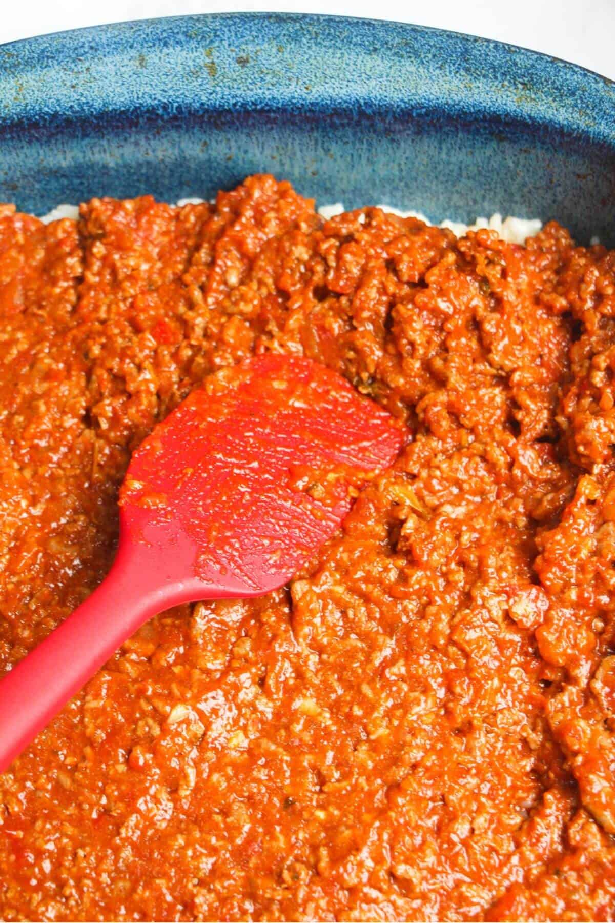 Red spatula pressing bolognese into a blue oven dish.