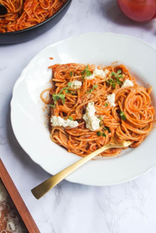 Bloody mary pasta, topped with burrata and basil, in a white scalloped bowl with more pasta in the background.