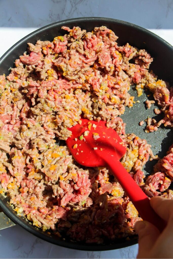 Red spatula stirring ground beef and browning it in a large pan.