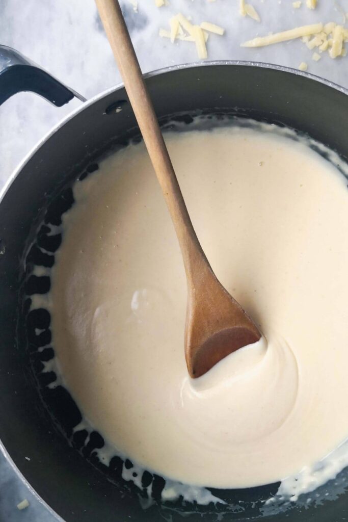 Cheesy bechamel sauce in a large black pot with a wooden spoon inside.
