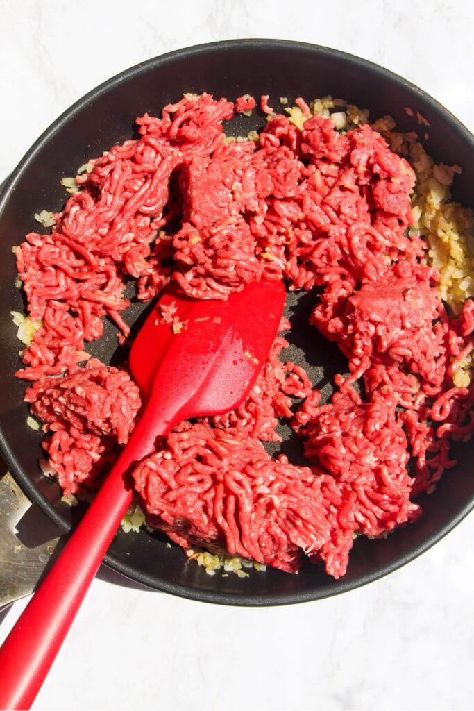 Ground beef with garlic and onions in a large black pan being stirred by a red spatula.