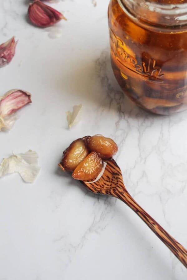 Garlic confit cloves on a wooden spoon on a white marble background, with a jar of garlic confit behind.