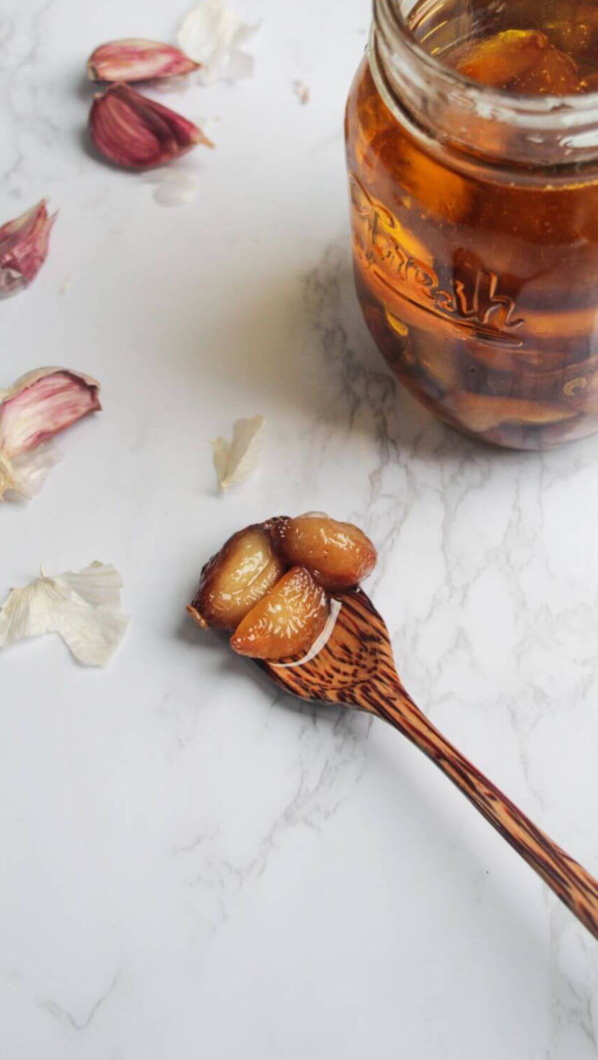 Garlic confit cloves on a wooden spoon on a white marble background, with a jar of garlic confit behind.