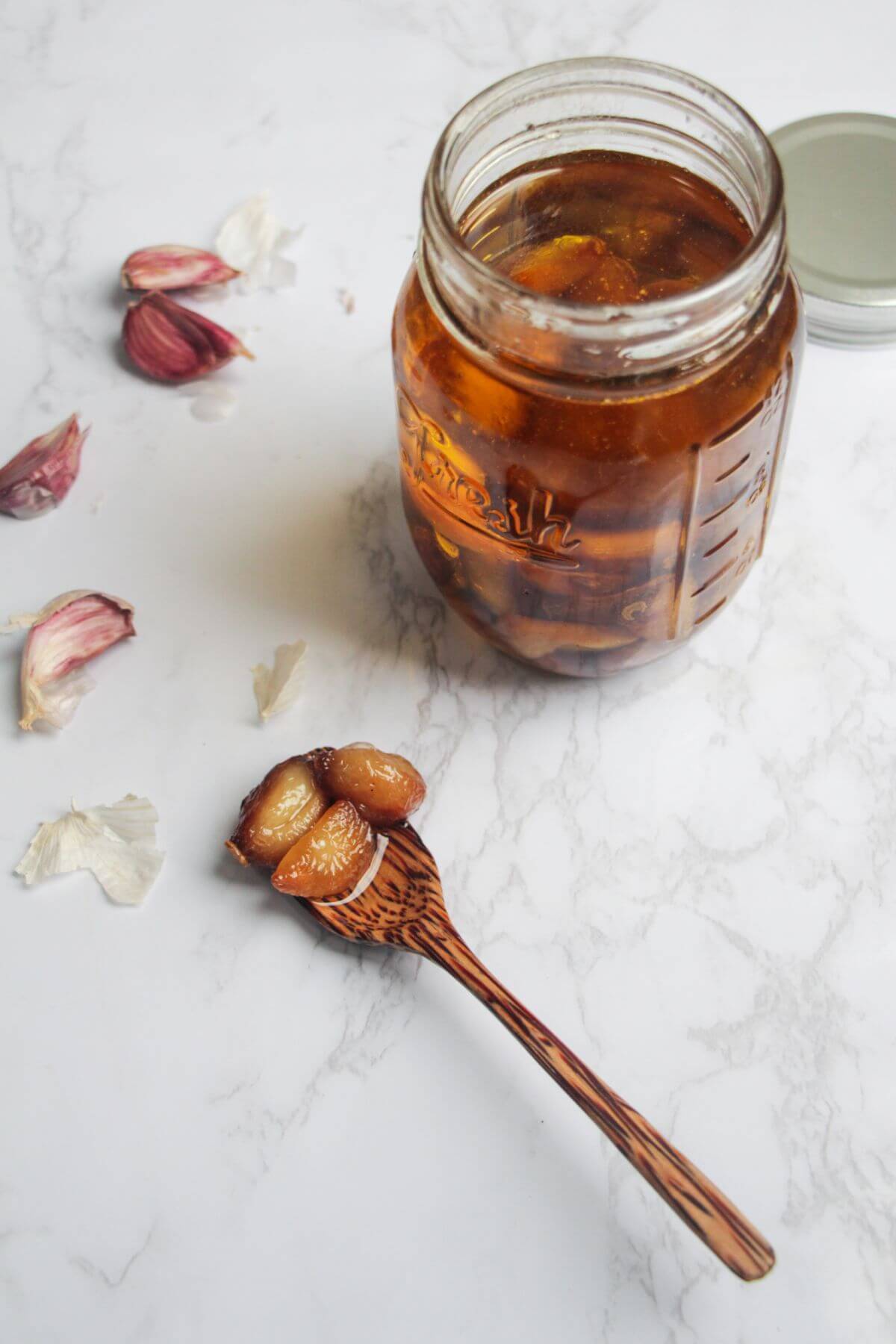 Garlic confit cloves on a wooden spoon on a white marble background, with a jar of garlic confit behind.