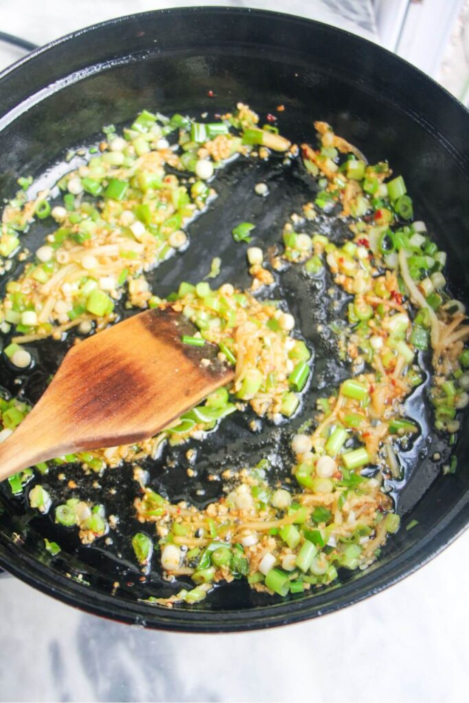 Wooden spoon stirring ginger, garlic and scallions in a large black pan.