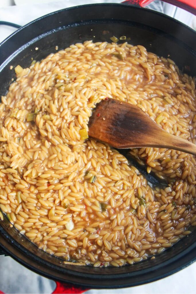 Wooden spoon stirring cooked orzo in a large black pan.