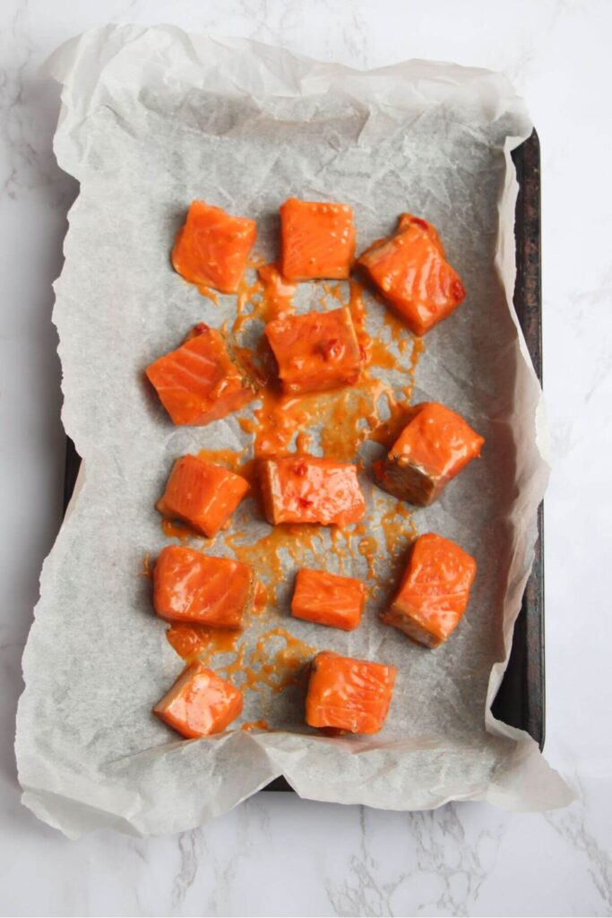 Diced salmon pieces tossed through bang bang sauce on a lined baking tray.
