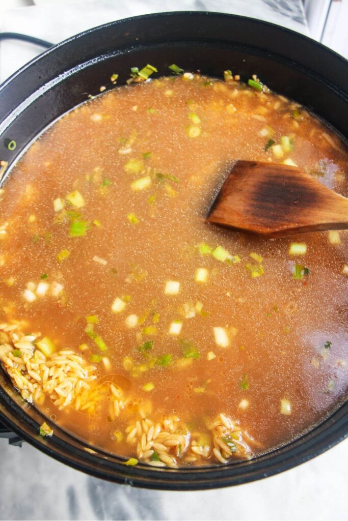 Chicken stock added to orzo, ginger, chilli, garlic and scallions in a large black pan.
