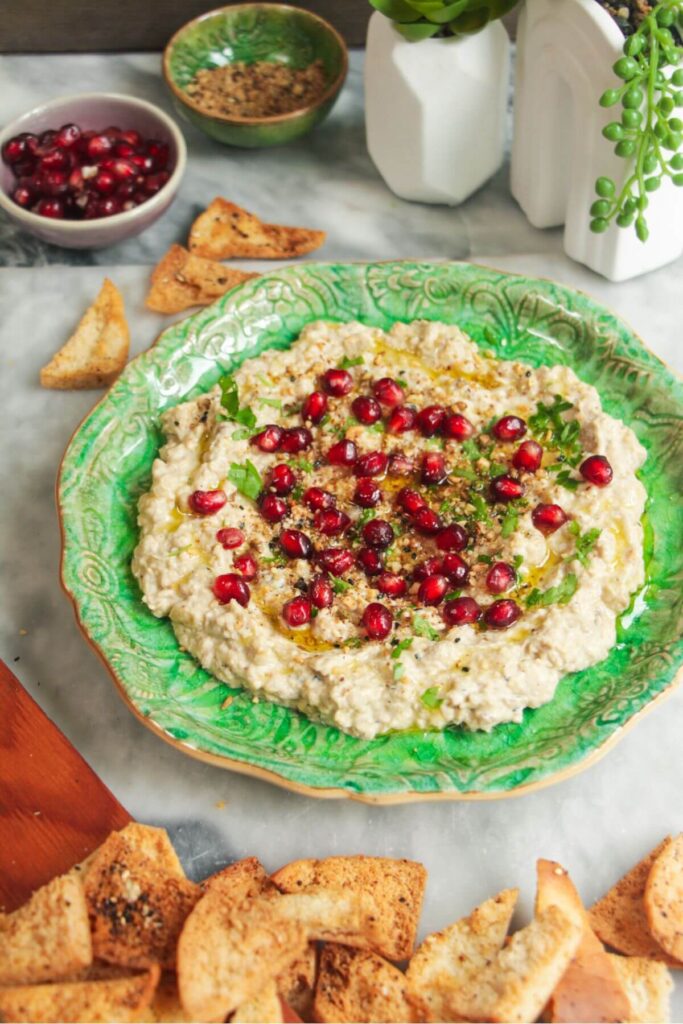 Smoky eggplant dip on a small green plate, with pomegranate seeds on top and pita chips on the side.