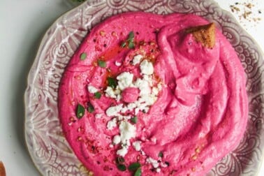 Bright pink beet feta dip on a pink plate with feta crumbled on top and pita chips on the side.