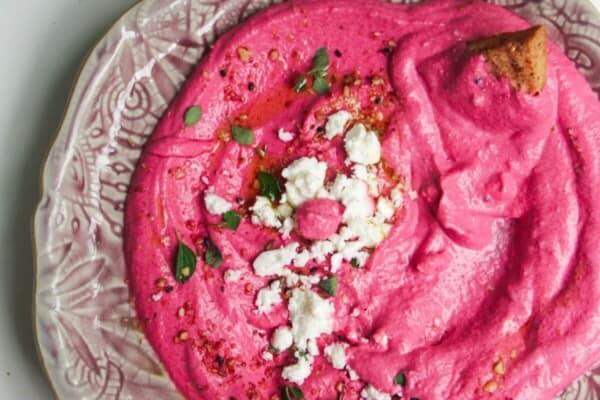 Bright pink beet feta dip on a pink plate with feta crumbled on top and pita chips on the side.