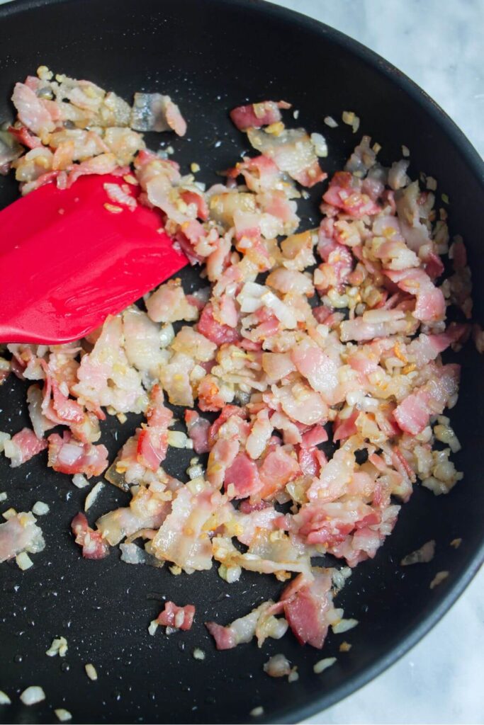 Diced bacon, onion and garlic cooking in a small black pan.