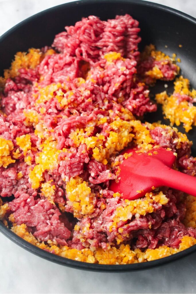 Red spatula stirring ground beef, carrot, celery, onion and garlic in a black pan.
