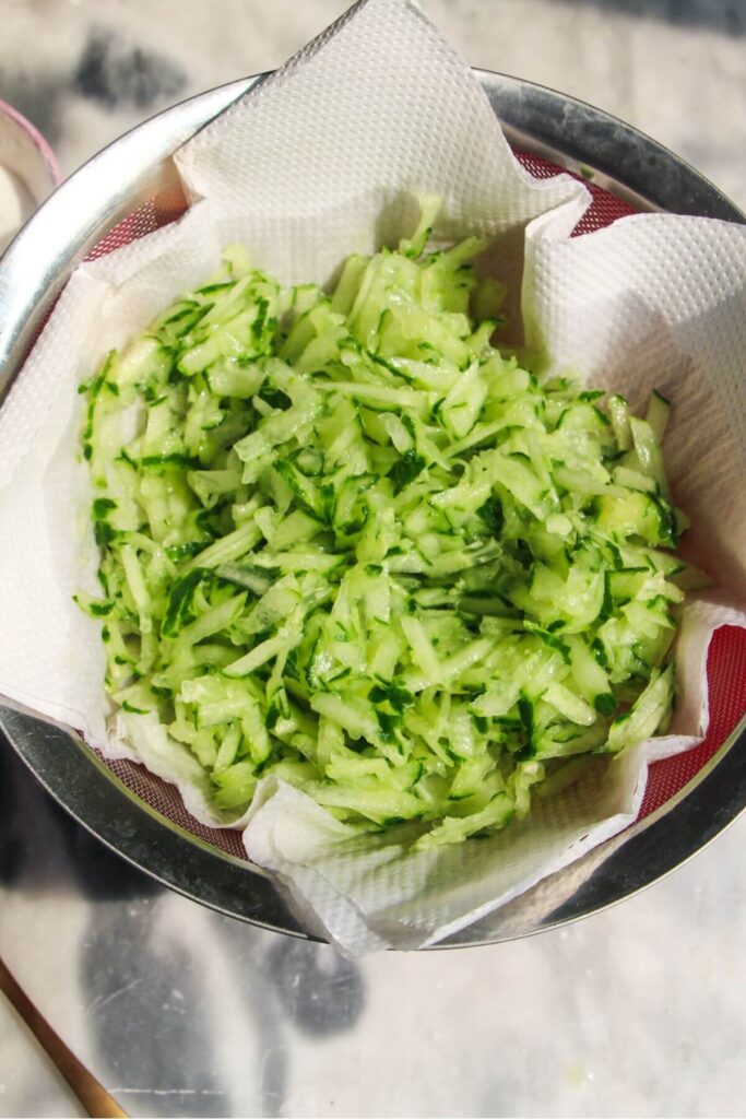 Grated cucumber in kitchen towel in a sieve set over a small pink bowl.