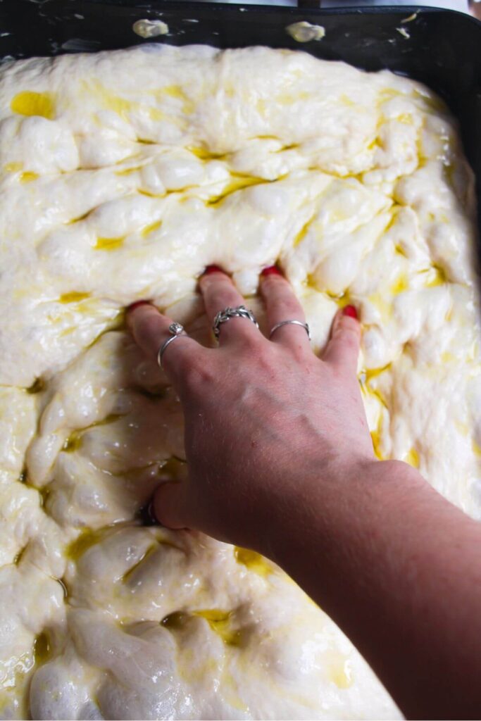 Fingers dimpling focaccia dough in a large oven pan.