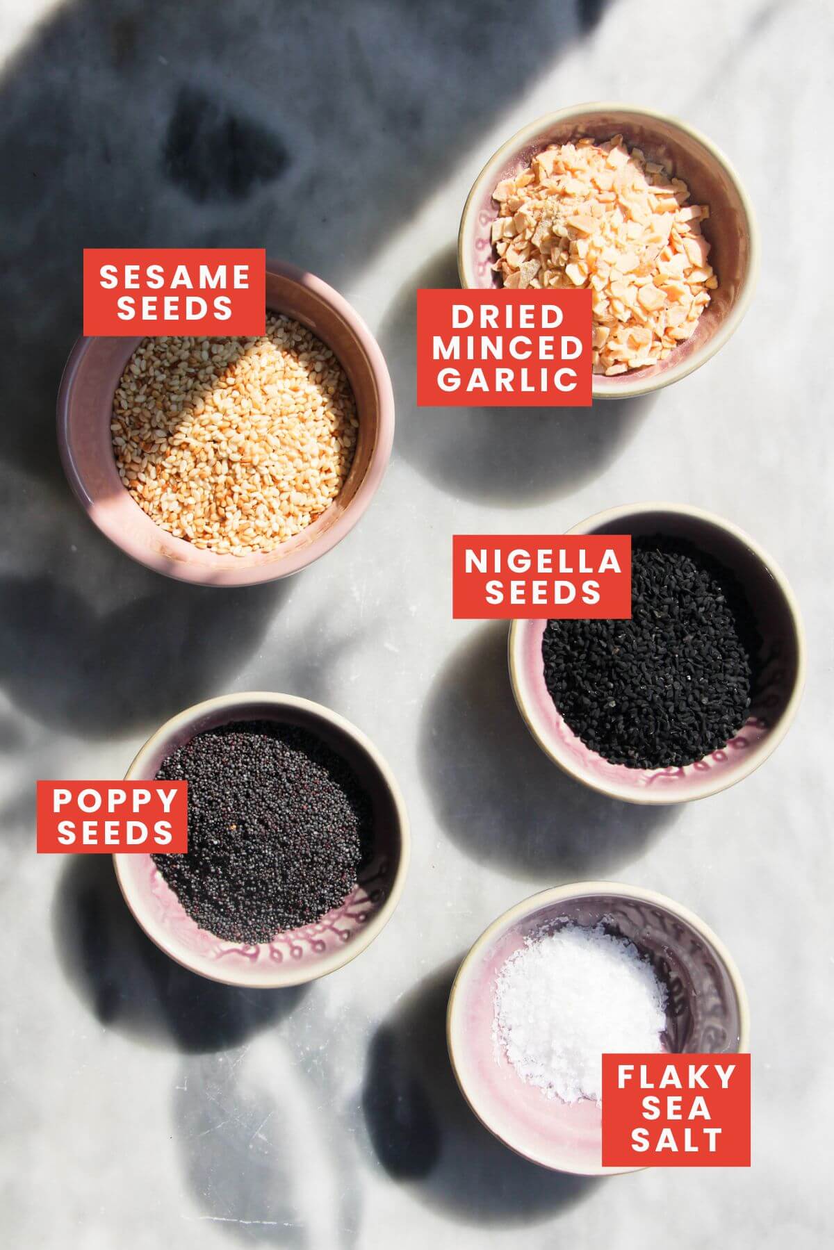 Ingredients for everything bagel seasoning laid out on a grey marble background and labelled.