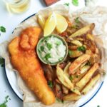 Beer battered fish fillet with a pile of chips and a small pot of tartar sauce in baking paper lined plate.