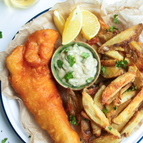 Beer battered fish fillet with a pile of chips and a small pot of tartar sauce in baking paper lined plate.