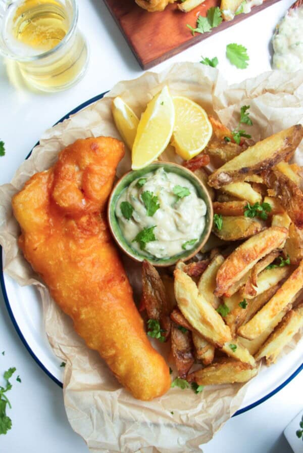 Beer battered fish fillet with a pile of chips and a small pot of tartar sauce in baking paper lined plate.