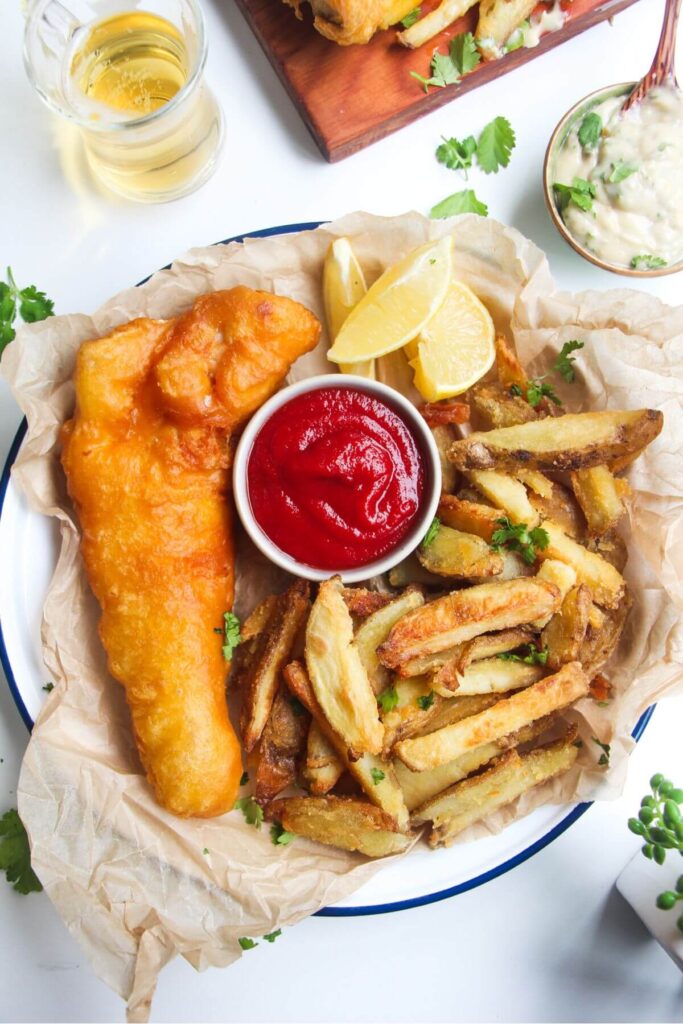 Beer battered fish fillet with a pile of chips and a small pot of tomato sauce in baking paper lined plate.