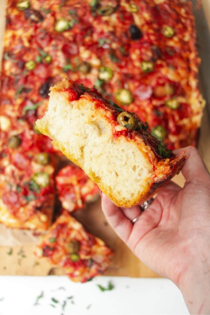 Side view of a hand holding up focaccia pizza, with the rest of the pizza in the background.