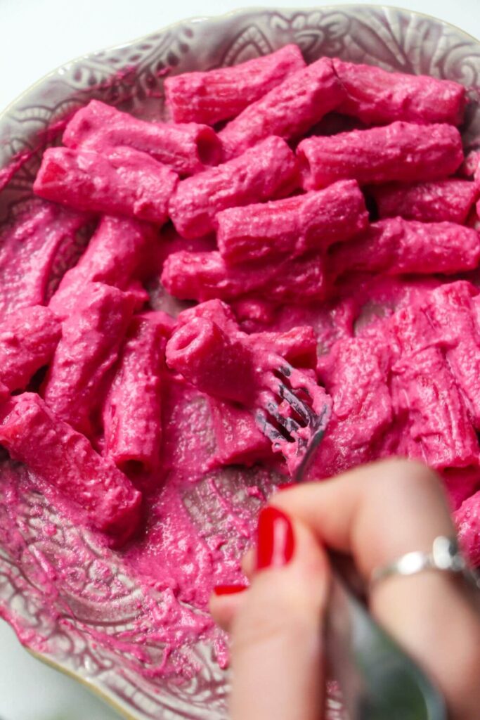 Close up of bright pink rigatoni pasta on a pink plate with a hand holding a fork.