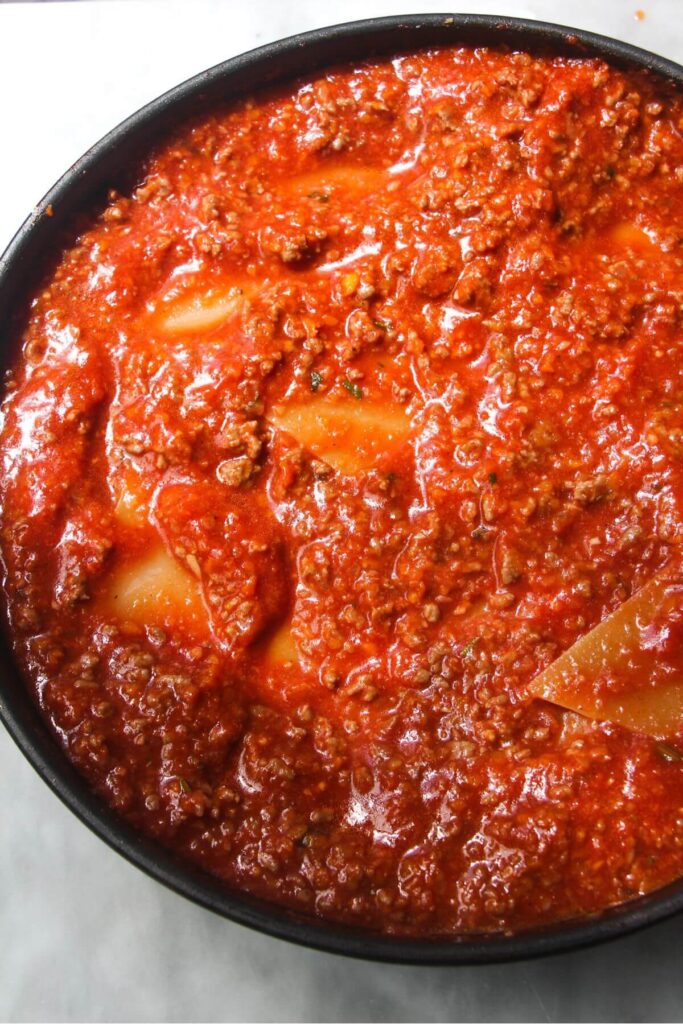 Lasagna sheets added into bolognese ground beef in a small black pan.