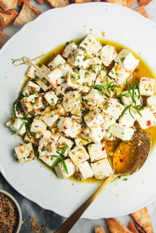 Marinated feta in a white scallop edges plate with a gold spoon inside and pita chips on the side.
