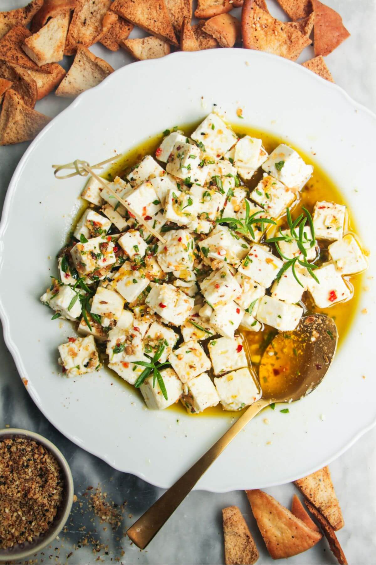 Marinated feta in a white scallop edges plate with a gold spoon inside and pita chips on the side.