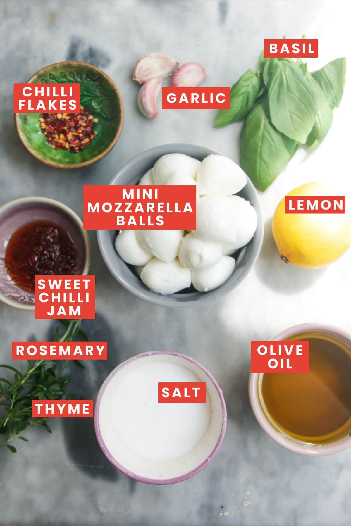 Ingredients for marinated mozzarella laid out on a grey marble background and labelled.