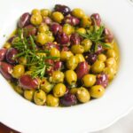 Close up of a white scalloped bowl of marinated olives, with skewers on the side.