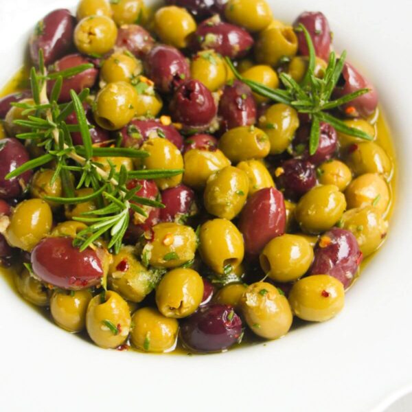 Close up of a white scalloped bowl of marinated olives, with skewers on the side.