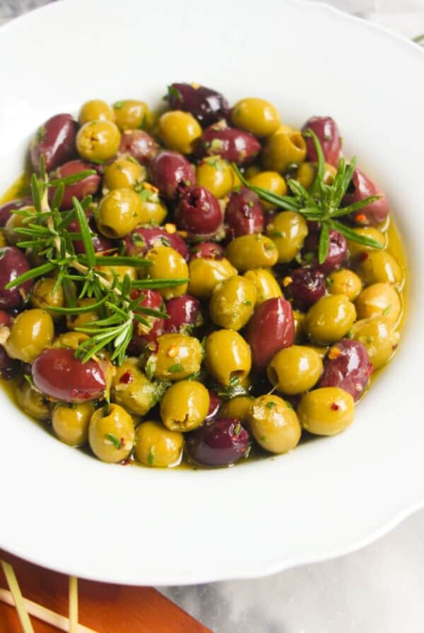 Close up of a white scalloped bowl of marinated olives, with skewers on the side.