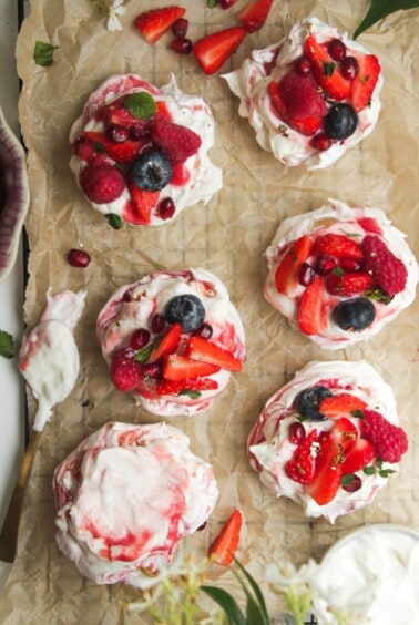 Six mini pavlovas on a baking paper lined tray topped with cheesecake cream and mixed berries.