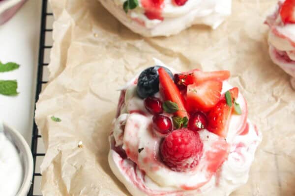 Two mini, berry topped pavlovas on a baking paper lined wire rack with more pavlovas behind them.