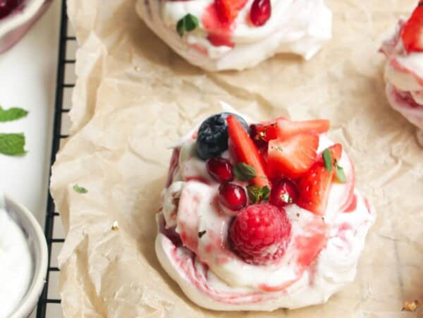Two mini, berry topped pavlovas on a baking paper lined wire rack with more pavlovas behind them.