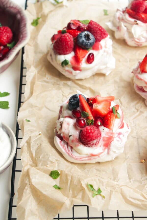 Two mini, berry topped pavlovas on a baking paper lined wire rack with more pavlovas behind them.