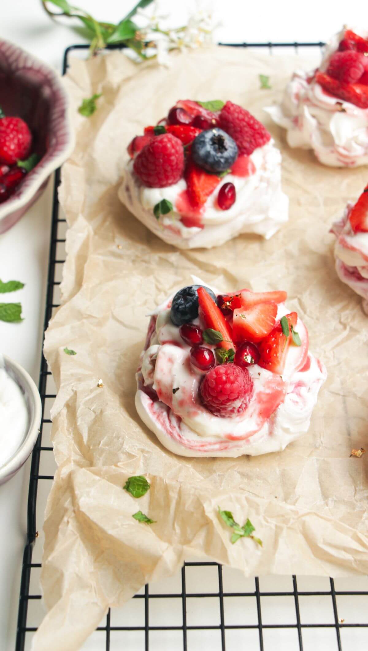 Two mini, berry topped pavlovas on a baking paper lined wire rack with more pavlovas behind them.