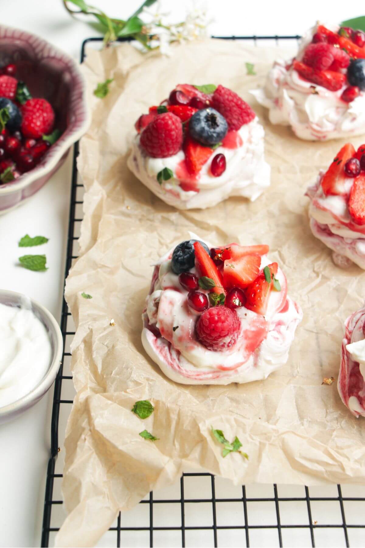 Two mini, berry topped pavlovas on a baking paper lined wire rack with more pavlovas behind them.