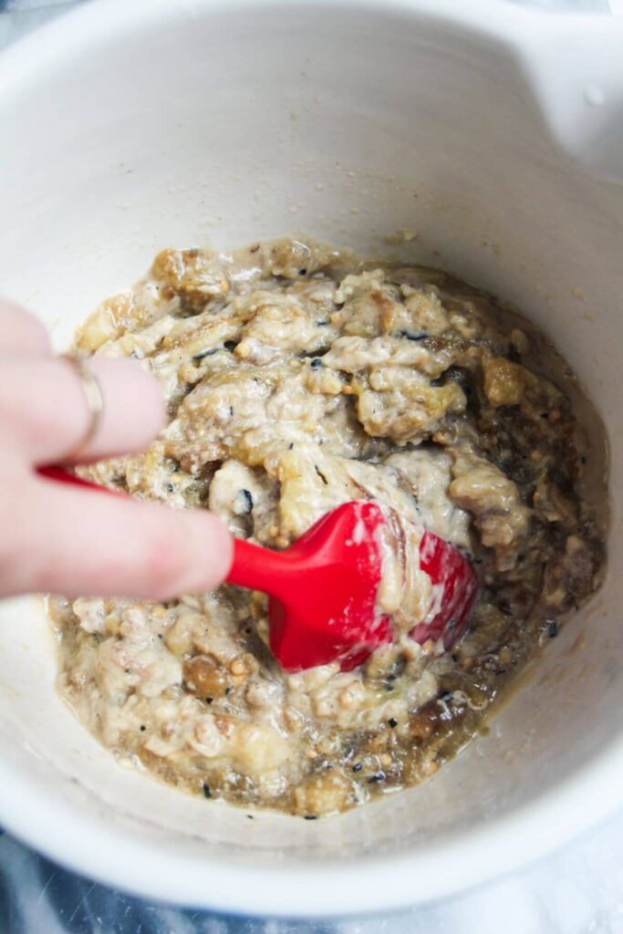 Hand holding a red spatula mixing smoky eggplant dip in a white bowl.