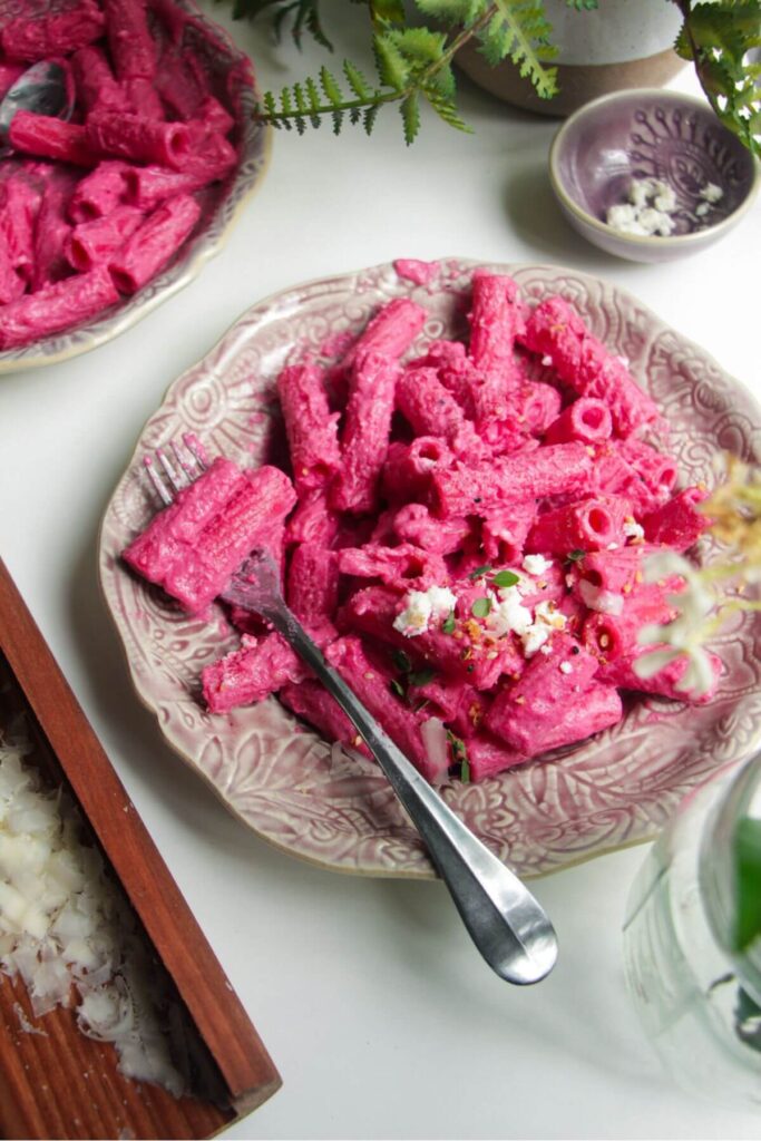 Bright pink pasta on a small pink plate, with a silver fork and another plate of pasta in the background.